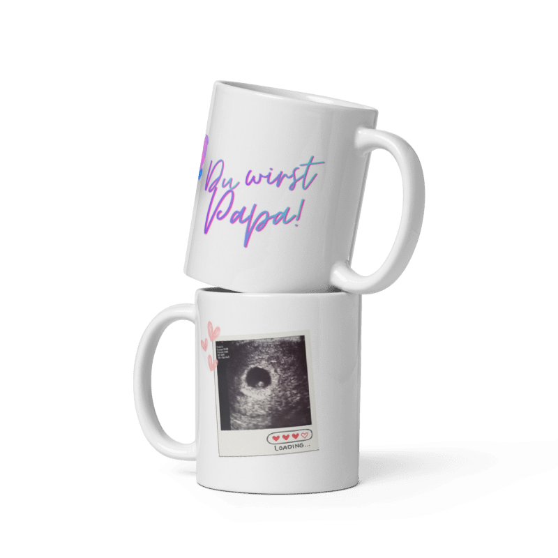 Zwei weiße Kaffeetassen übereinander gestapelt: Die obere zeigt den geschwungenen, blau-violetten Schriftzug „Du wirst Papa!“, die untere ein echtes Ultraschallbild im Polaroid-Design mit kleinen rosa Herzen und der Aufschrift „Loading...“.