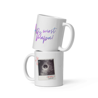 Zwei weiße Kaffeetassen übereinander gestapelt: Die obere zeigt den geschwungenen, blau-violetten Schriftzug „Du wirst Papa!“, die untere ein echtes Ultraschallbild im Polaroid-Design mit kleinen rosa Herzen und der Aufschrift „Loading...“.