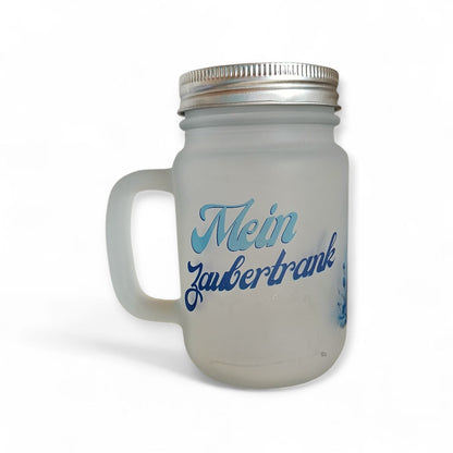 Ein elegantes, mattiertes Trinkglas mit Henkel und silberfarbenem Schraubdeckel, das mittig mit dem Schriftzug „Mein Zaubertrank“ in blauen Buchstaben bedruckt ist.