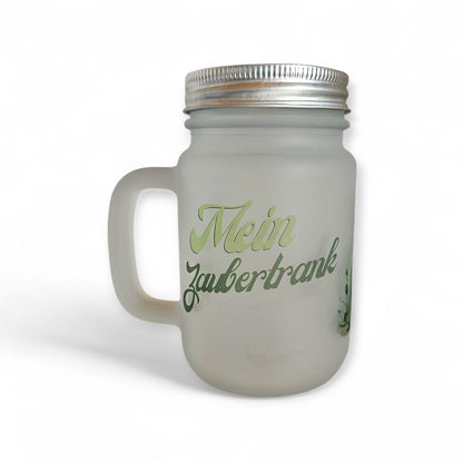 Ein elegantes, mattiertes Trinkglas mit Henkel und silberfarbenem Schraubdeckel, das mittig mit dem Schriftzug „Mein Zaubertrank“ in gruenen Buchstaben bedruckt ist.