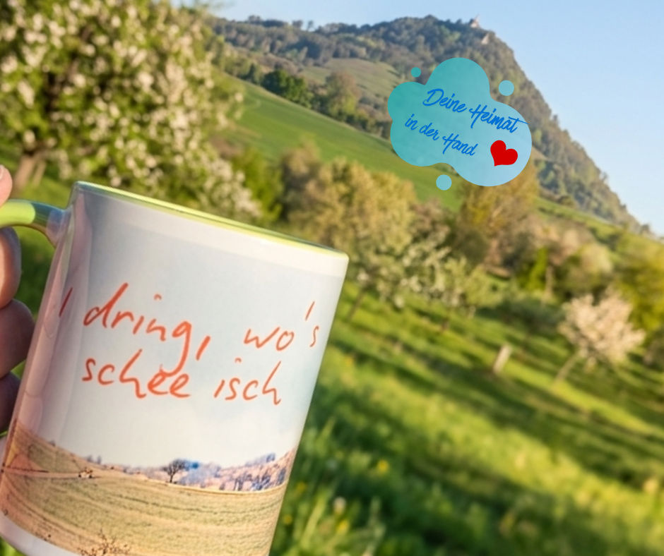 Eine Nahaufnahme einer personalisierten Schlucksachen-Tasse mit dem schwäbischen Spruch „I dring, wo’s schee isch“. Im Hintergrund die blühende Landschaft und die Achalm in Eningen unter Achalm. Eine grafische Wolke im Himmel enthält den Text „Deine Heimat in der Hand ❤️“.