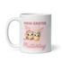 Weiße Keramiktasse mit dem Motiv Mein erster Muttertag, abgebildet mit einer Kaffeetasse, einem Babyfläschchen und kleinen Herzen, Henkel auf der linken Seite.